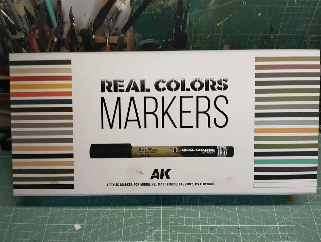 Boîte de marqueurs REAL COLORS de AK, avec une large gamme de couleurs, incluant un marqueur RCM018 RAL 7028 DUNKELGELB. Indiqué comme marqueurs acryliques pour modélisme, finition mate, séchage rapide et imperméable. Divers outils de modélisme en arrière-plan."