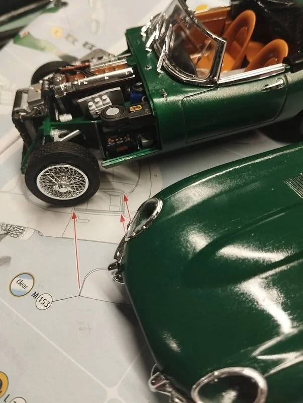 Maquette de voiture verte en cours d'assemblage, montrant les détails du capot et des roues avec des flèches rouges indiquant les points de fixation.